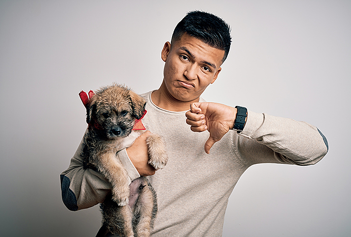 man grimaces and puts a thumbs down hand gesture while holding a small brown dog