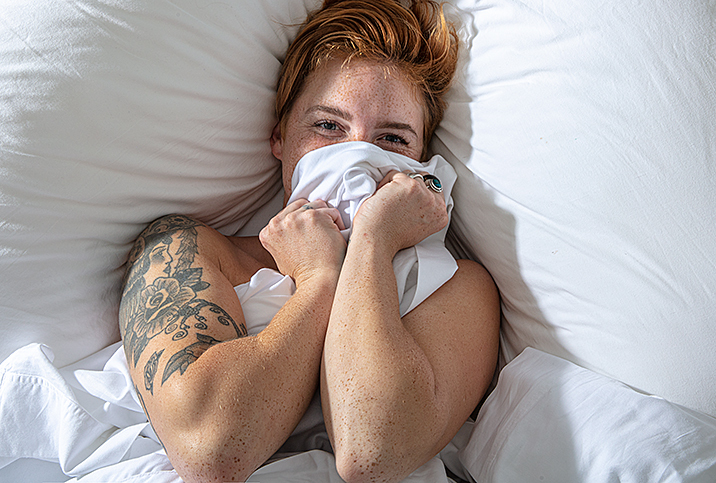 woman with tattoos lays in bed with sheets in hand covering her mouth