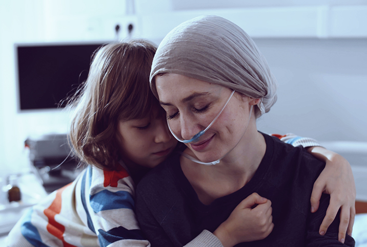 A young child hugs their mother who is wearing a head scarf and oxygen cannula.