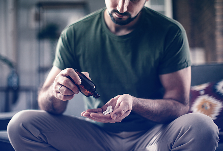 A man pours white pills from a brown bottle into his hand while sitting down.
