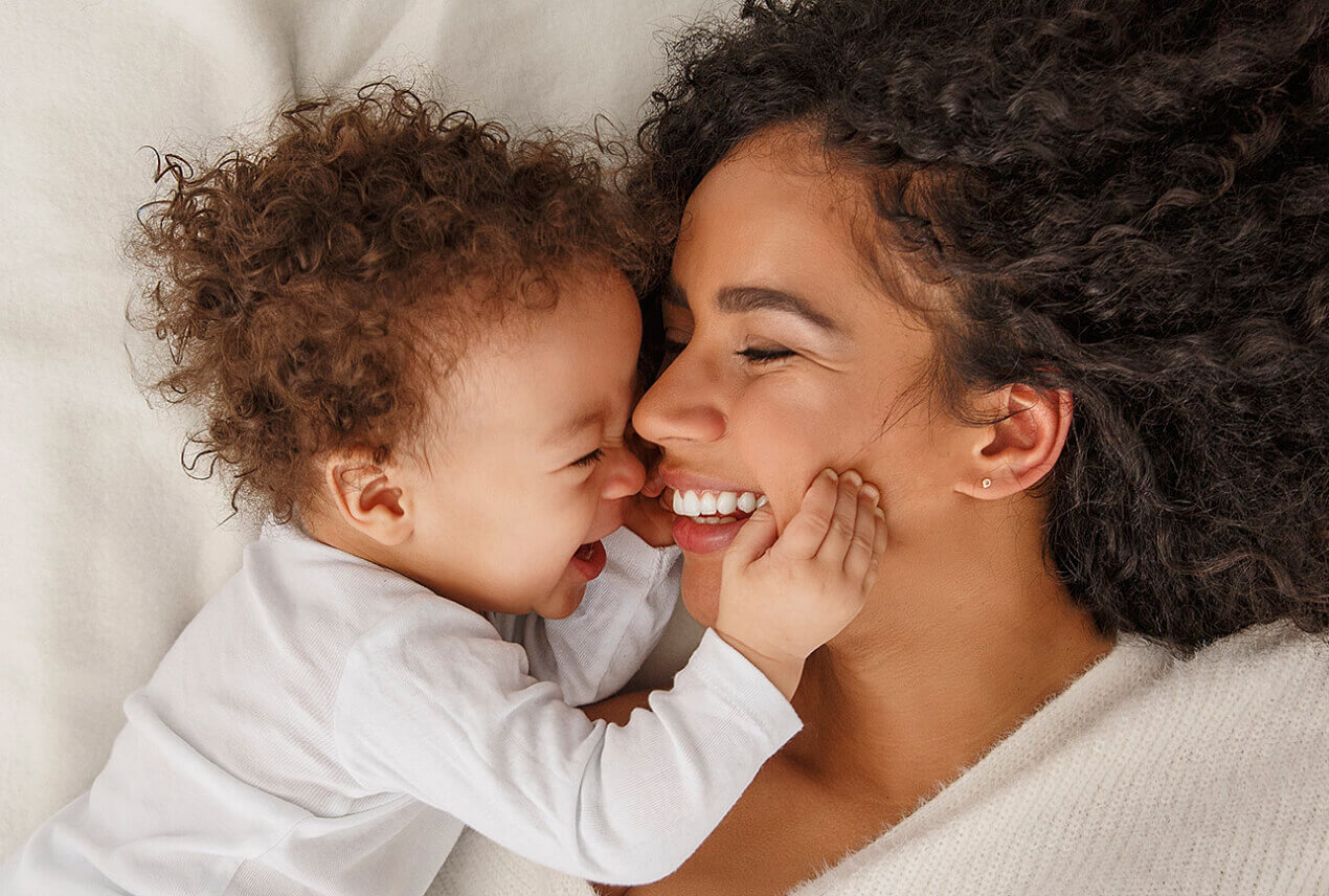 A mother and her baby face each other smiling in bed.