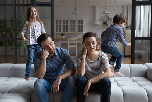 A couple sits on a sofa as their kids play around them.