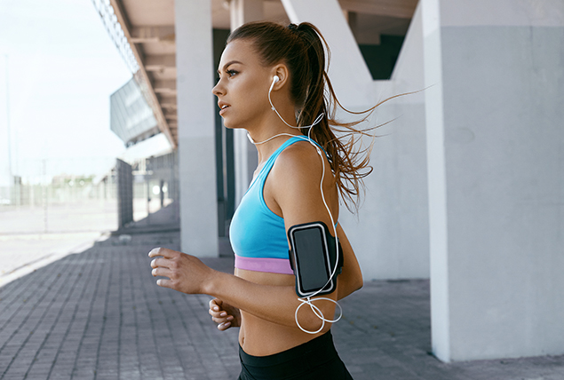 A woman is running outside with an iphone attached to her arm and wired earbuds in.