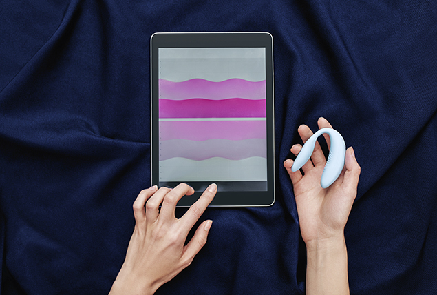 hands hold white vibrator and tap on tablet with pink ruffled graphic on navy sheets