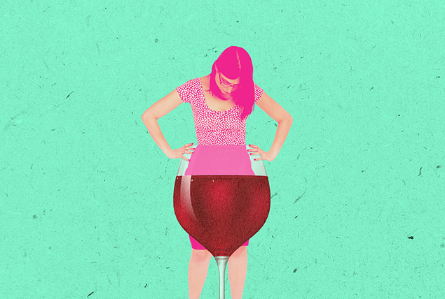 A woman stands behind a large glass of wine looking down towards her abdomen.
