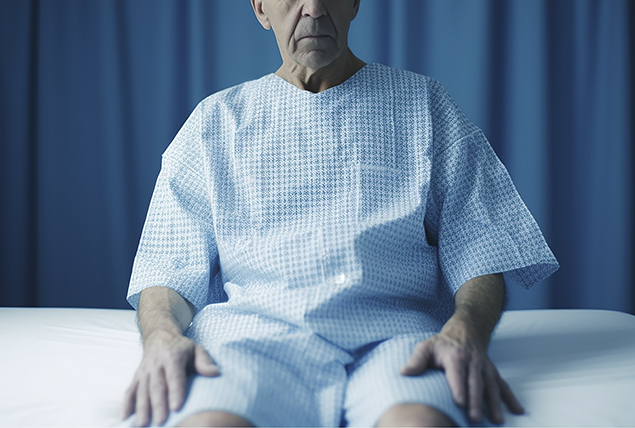 An older man sits on a medical bed in a hospital gown.