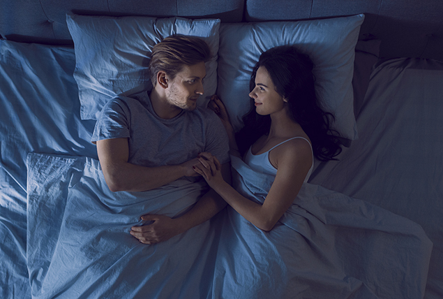 man and woman lay in bed together in darken room, holding hands and smiling at each other