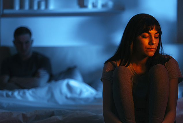 A woman sits on the end of a bed while a man sits behind against the headboard.