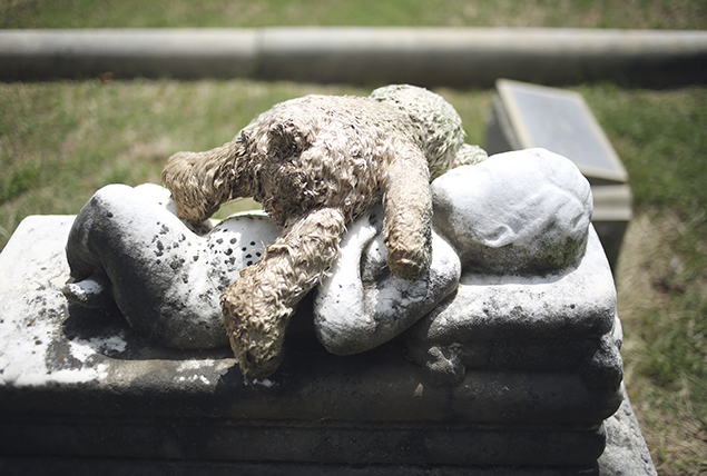 A teddy bear lays on top of a gravestone with an infant engraved on it.