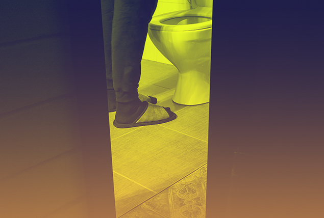 A pair of legs in slippers stand in front of a toilet in a dimly lit bathroom.