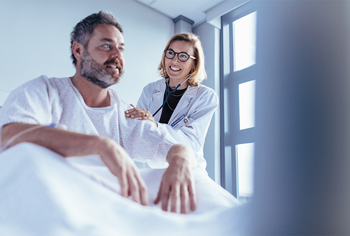 A man sits up in a hospital bed while a doctor listens to his back with a stethoscope. 