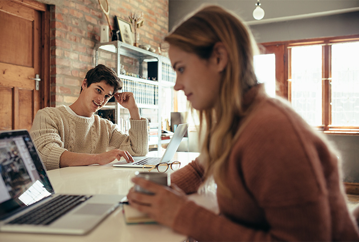 A man looks over at his partner while both are sitting at a table together on their laptops.