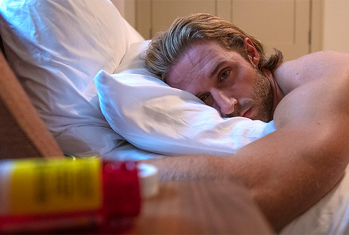 A man lays on his stomach on a bed with his head facing the side and looking sad.