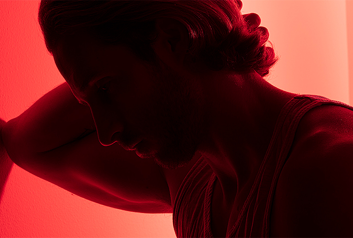 A man leans against a wall while holding his head with a red light shining behind him.