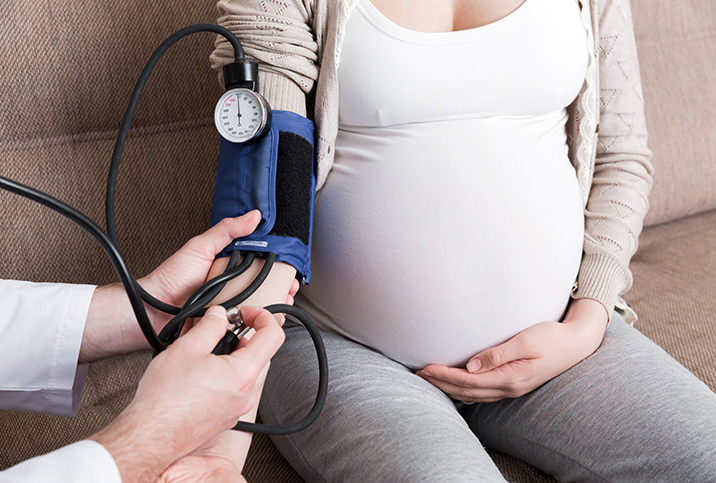 A pregnant woman holds her belly while having her blood pressure tested by a doctor.