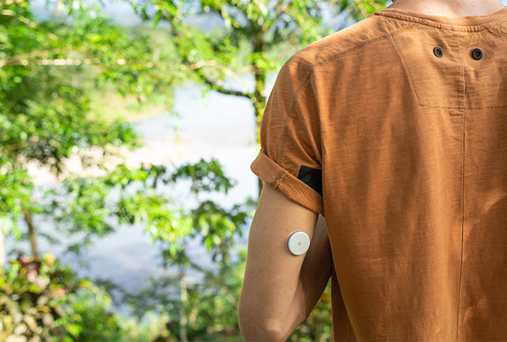 A man with diabetes stands near trees with his back to the camera with a glucose meter on his arm.