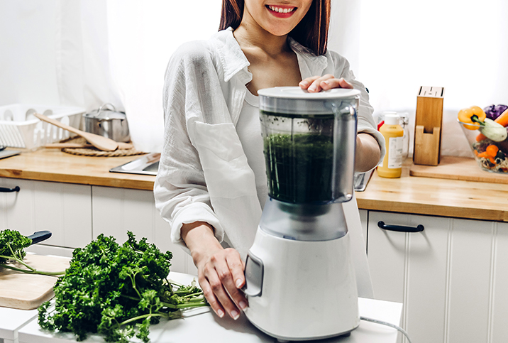 A smiling woman in a kitchen stands next to a cutting board with parsley on it and places her hands on a blender.