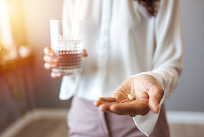 A person holds a glass of water in their right hand, and two pills in their left hand, which is in the foreground.