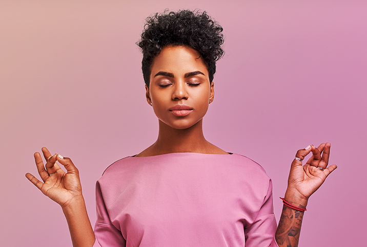 A woman in a pink shirt meditates with her eyes closed against a pink background.