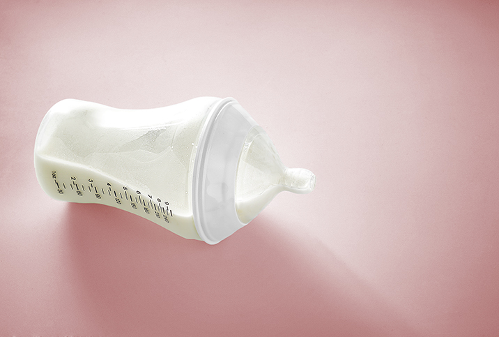 A baby bottle with milk lays on its side against a pink background.