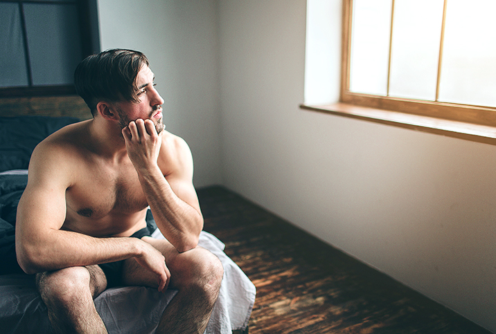 A man sits on the end of his bed scratching his beard and looking out the window.