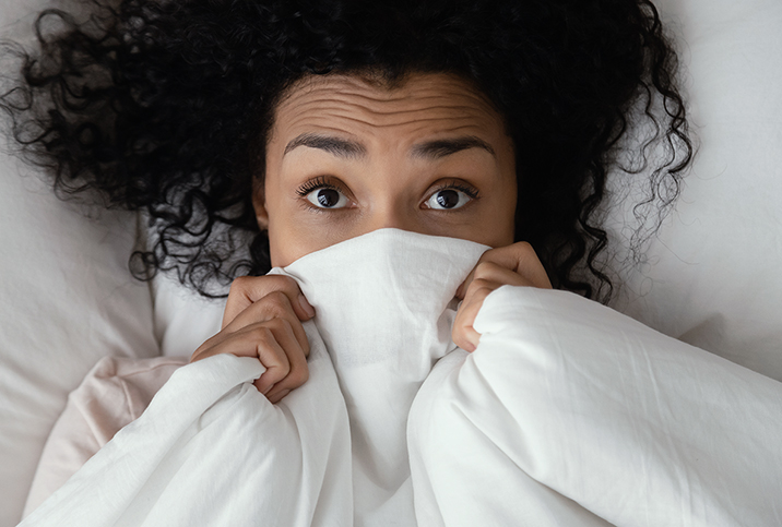 A woman lies in bed with a blanket over the lower part of her face with her eyes wide in fear.