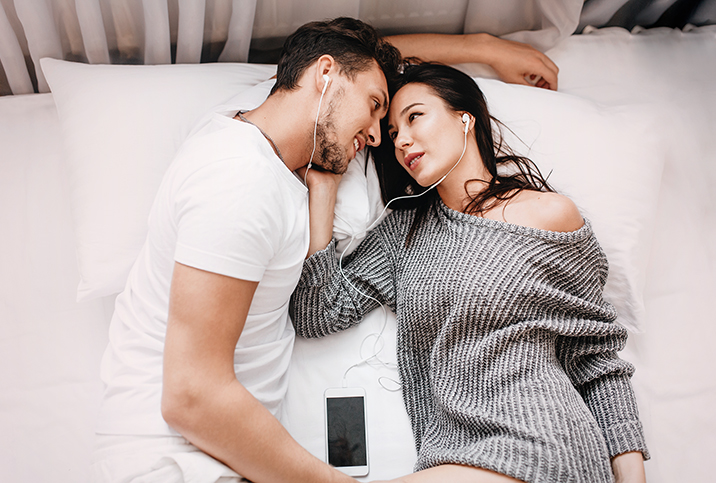 A couple lays in bed looking at each other sharing earbuds.