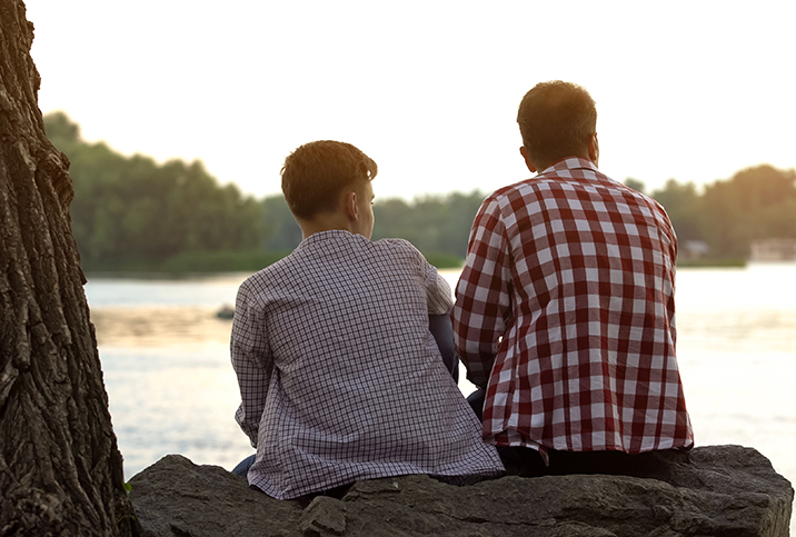 A father and son sitting on a rock face away from the camera and look at a river in front of them.