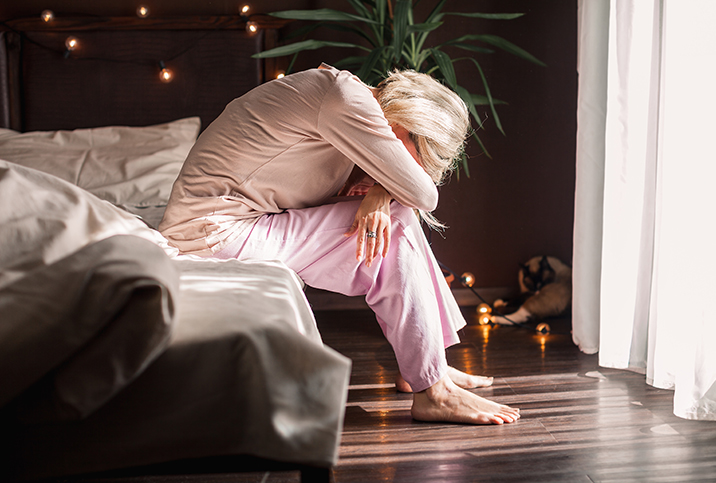 A woman sits on the edge of a bed with her feet on the ground and folds over her knees, resting her forehead on her folded arms.