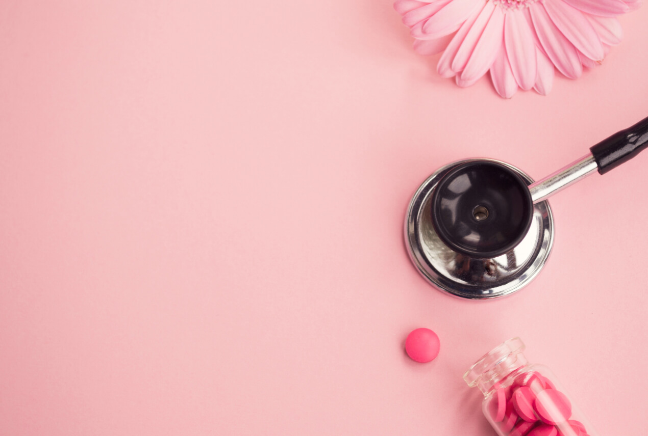 A pink pill spills out of a clear bottle onto a light pink surface next to a stethoscope.