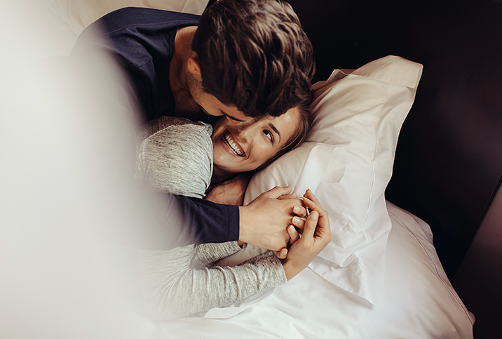 A couple cuddle in bed while smiling at each other.