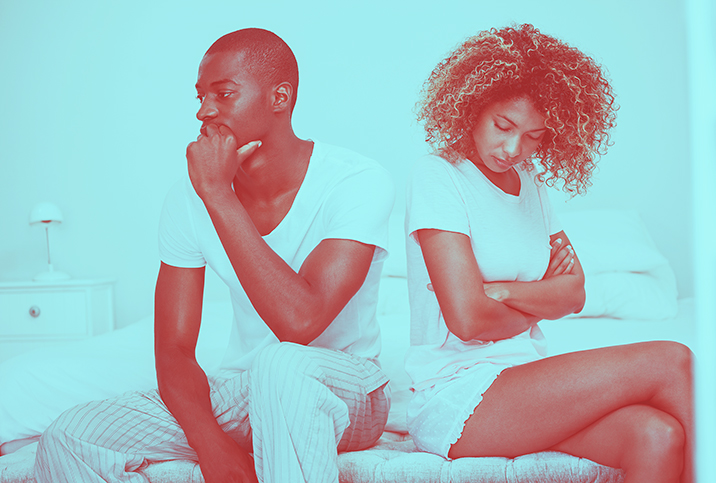 A couple sits on the edge of a bed facing away from each other and looking concerned.