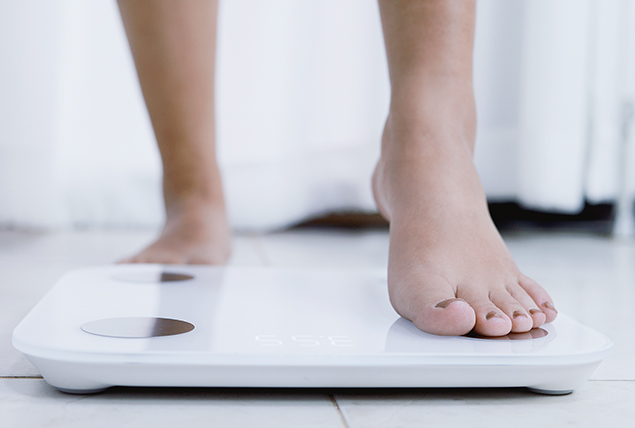 A person steps one foot onto a white scale as it sits on a white-tiled floor.