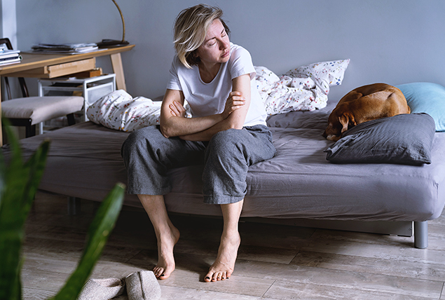 A woman sits on her bed with her eyes closed and her arms crossed against her knees.