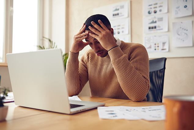 A man sits at his laptop holding his hands against the top of his head.