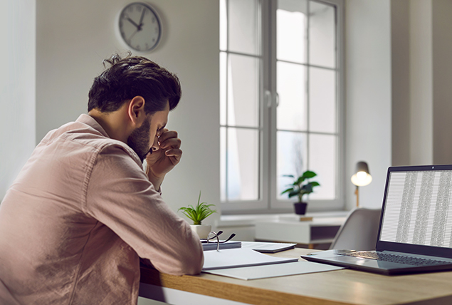 A man sits at a desk in front of papers and his laptop rubbing his nose.