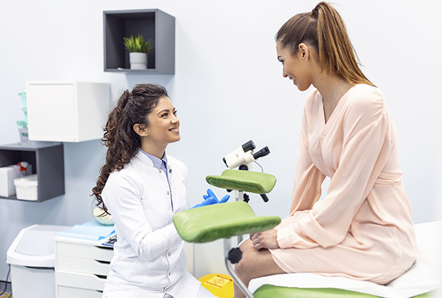 woman in pink shirt talks to doctor while sitting on pap smear bed