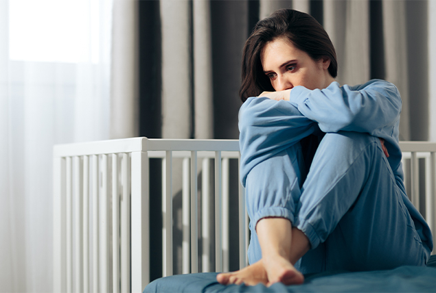 A woman sits on a bed with her knees curled up under her arms.