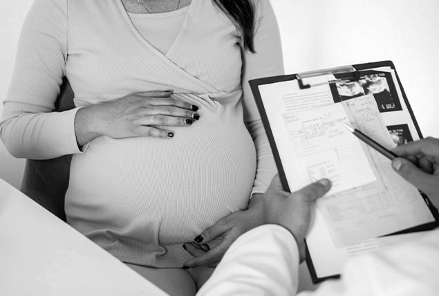 A pregnant woman holds her belly while listening to a doctor with a clipboard.