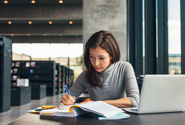 student writes on paper with blue pen with laptop open in college library