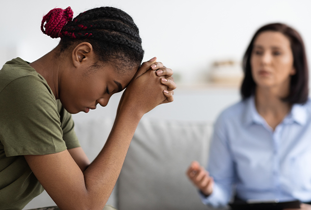 A woman sits in a chair with her head against her hands while another woman speaks to her.