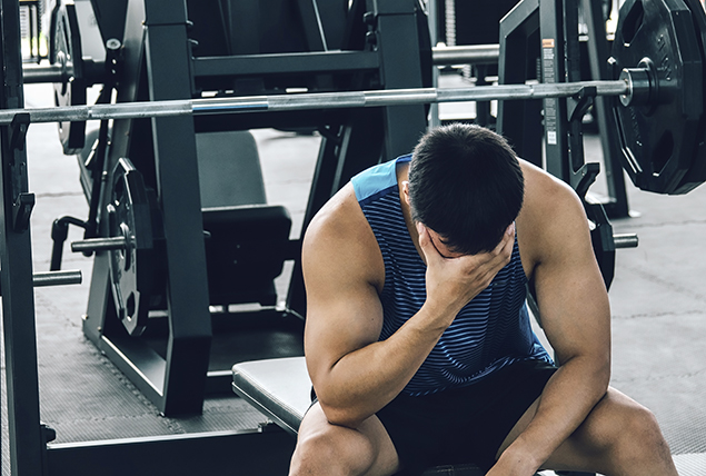 A man sits on a weight bench leaning over with his head in hand and elbows on his knees.