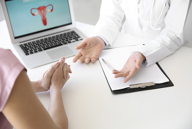 A doctor talks to a woman across a desk with a laptop in the backgroun.