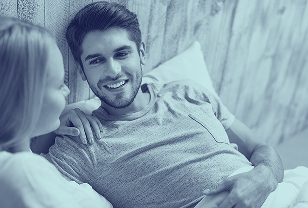 A new couple sits in bed smiling and talking to each other.