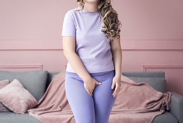 A woman in a grey t-shirt and jeans holds her hand over her groin area.