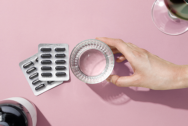 Two packets of activated charcoal pills are sat on a pink surface next to a glass of water.