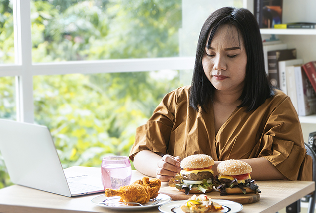 A woman sits in front of her laptop with two cheeseburgers and chicken wings in front of her.